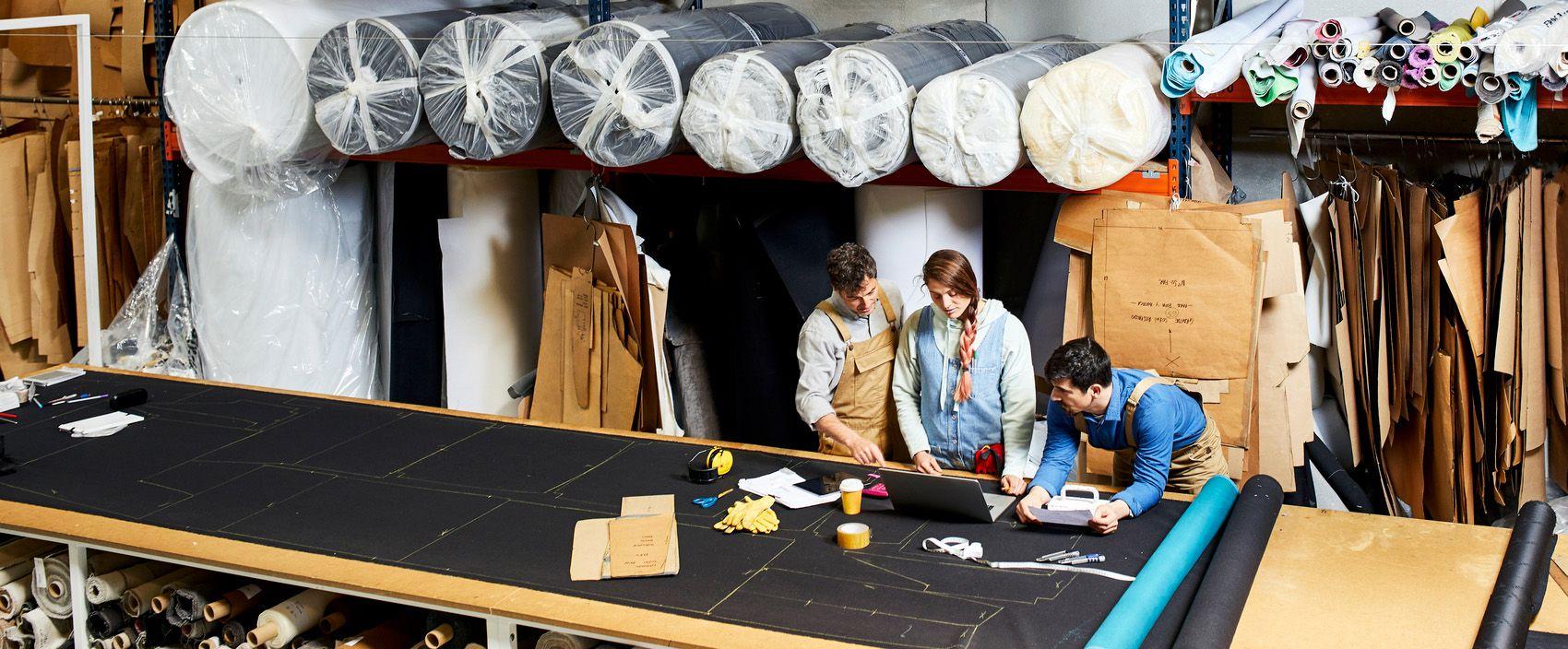 Drie mensen staan aan een ontwerptafel in een stoffenfabriek en hebben werkoverleg.