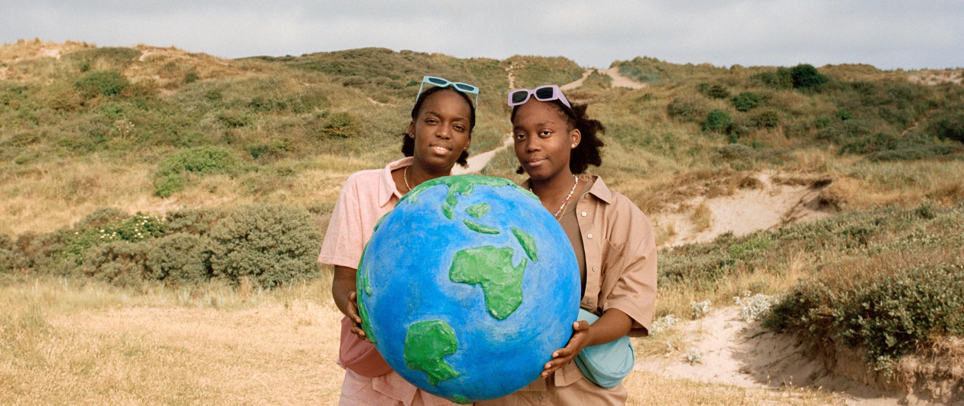 Twee meisjes staan in de duinen en houden een wereldbol vast. Ze kijken beiden in de camera.